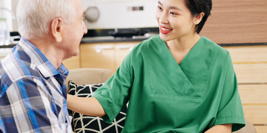 Positive young Asia female caregiver talking to senior patient and discussing his health and needs when visiting his at home